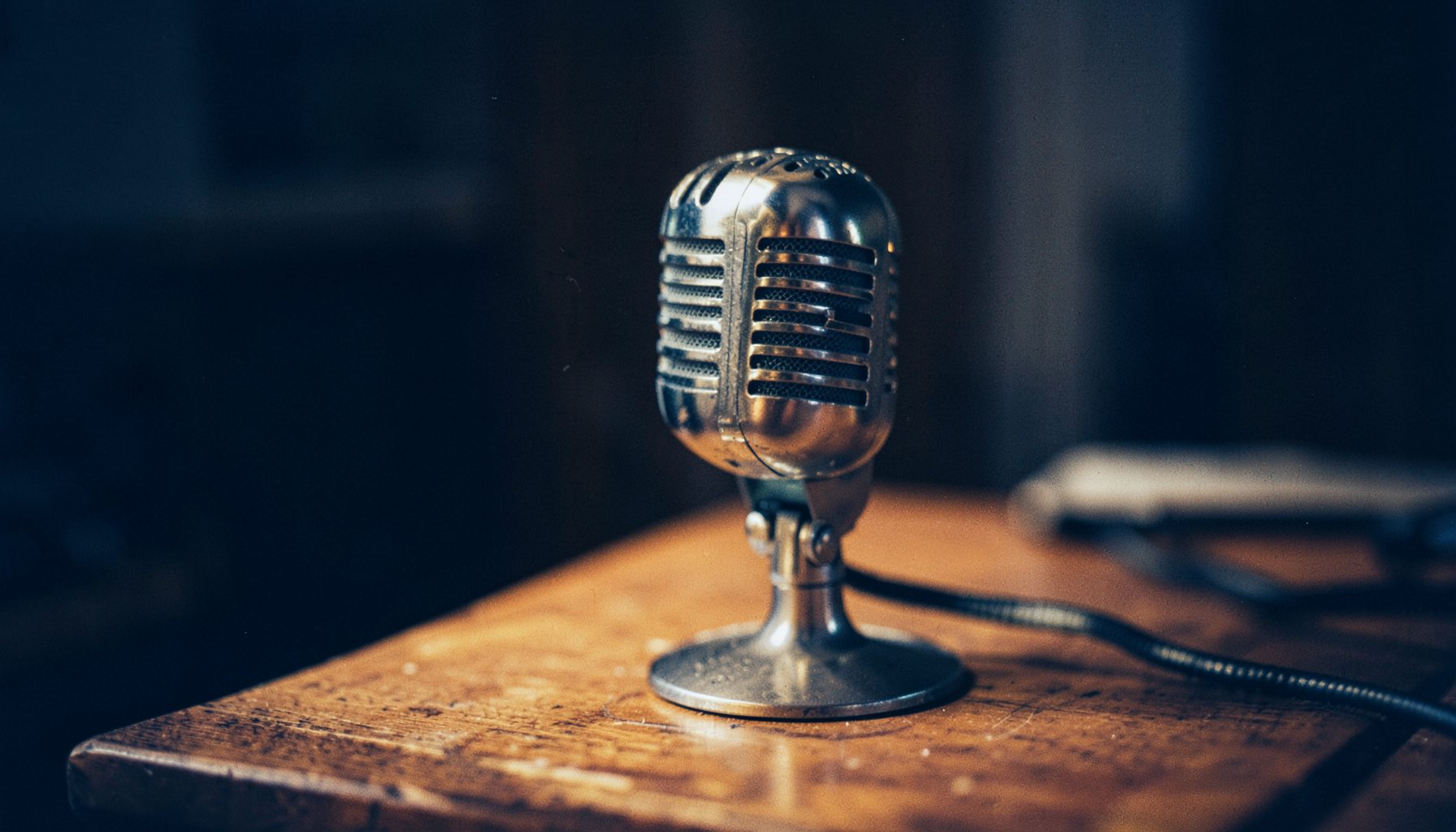 A warm-toned photograph of a vintage radio studio microphone on a wooden table, with soft amber lighting and a dark, moody background.