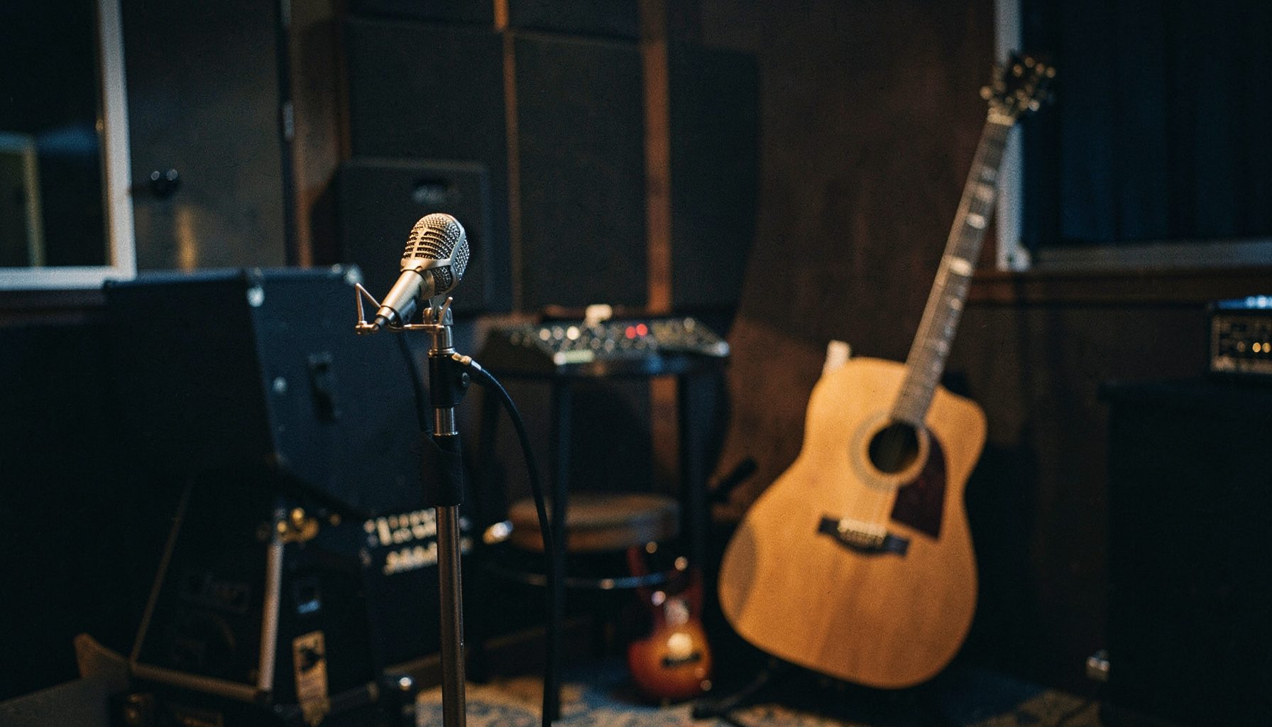 A warm, dimly lit recording studio with a vintage microphone on a stand in the foreground, acoustic guitar leaning against a stool, deep amber and brown tones